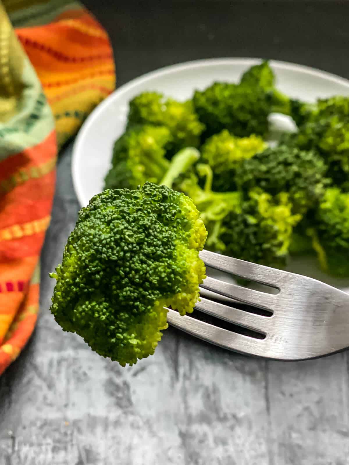A silver fork with a broccoli floret and a plate of steamed broccoli in the back.