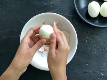 A pair of hands peeling the hard boiled egg over a white bowl.