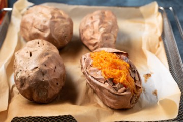 A parchment paper lined air fryer tray with four sweet potatoes and one cut open in the bottom right.
