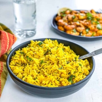 A black bowl filled with Indian Yellow Rice with a silver spoon on the right side and a plate of shrimp in the back.