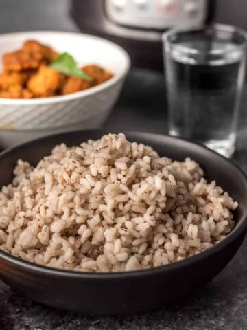 A black bowl with kreala matta rice after cooking in the Instant Pot, a cup of water in the back right, and a white bowl in the back left.