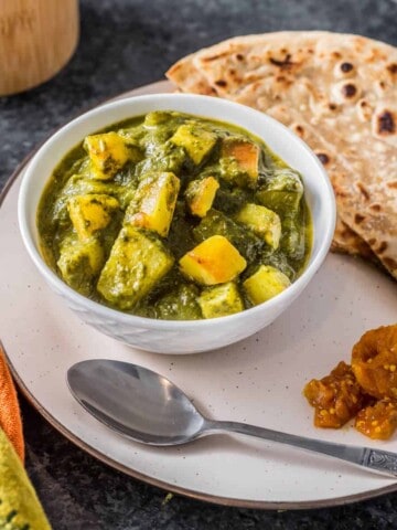 A white plate with a white bowl filled with aloo saag on the left of the plate and a silver spoon at the front of the plate with fresh paratha in the back of the plate.