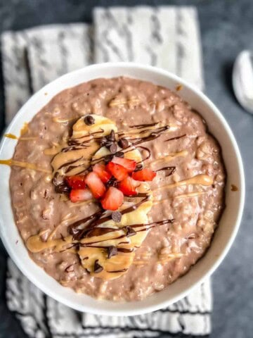 A white bowl filled with chocolate peanut butter oatmeal and topped with sliced banana, chopped strawberry, and a drizzle of peanut butter and chocolate with a spoon to the right of the bowl.