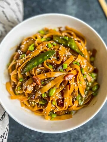 A white bowl with Instant Pot lo mein topped with sesame seeds with a white and black towel to the left and a set of chopsticks to the right.