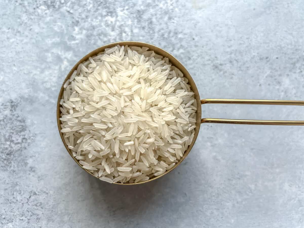 A gold measuring cup filled with uncooked Jasmine rice on a grey counter.