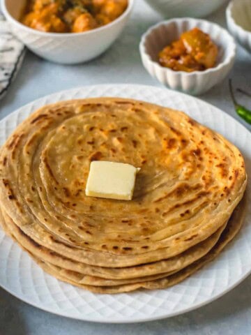 A white plate with a stack of lacha paratha topped with a pat of butter.