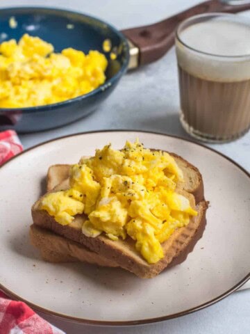 A white plate with a brown rim and a slice of toast topped with fresh scrambled eggs with a skillet in the back left and a glass of tea in the top right.