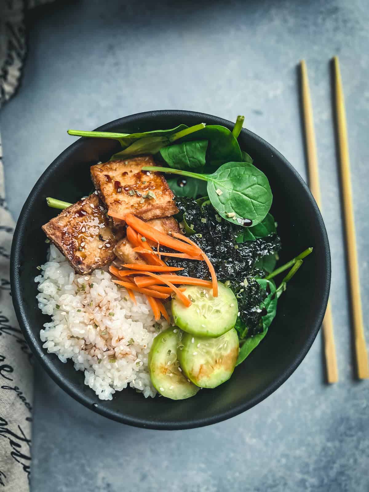 Top down black bowl with pressure cooker sushi rice cooked in the Instant Pot loaded with toppings. The toppings are seared glazed tofu, shredded carrots, sliced cucumbers, nori, and baby spinach.