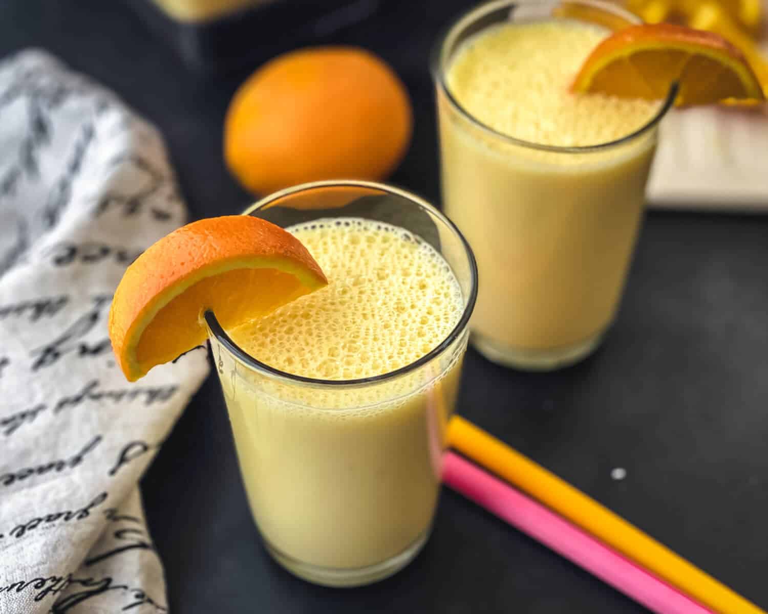 Two glasses of orange smoothie with an orange slice on the rim, two straws in between, and an orange in the back.