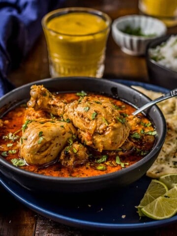 A close up shot of Indian curry chicken served in a gray bowl accompanied with naan and lassi