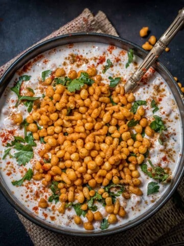 An overshot of boondi raita garnished with cilantro and served in a black bowl with a golden spoon.